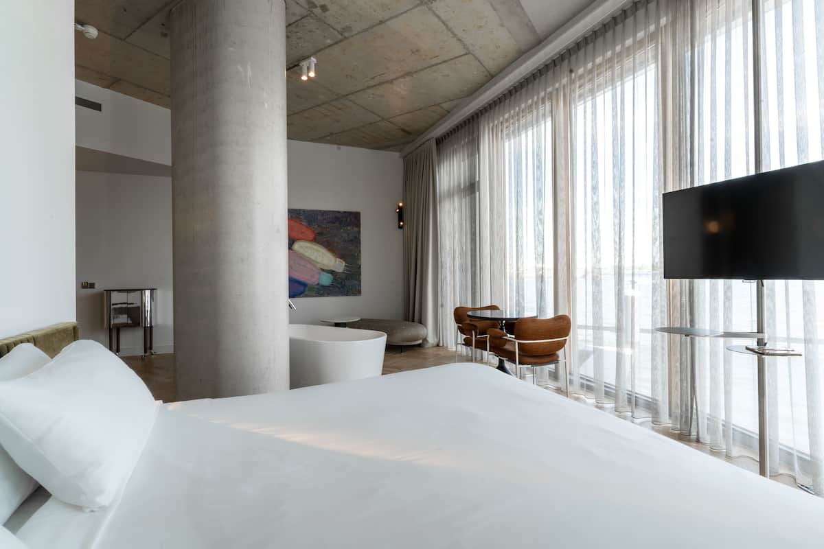 Moderni hotellihuone, jossa on suuri sänky, taulutelevisio, oleskelualue tuoleineen sekä suuri ikkuna, jossa on verhot.