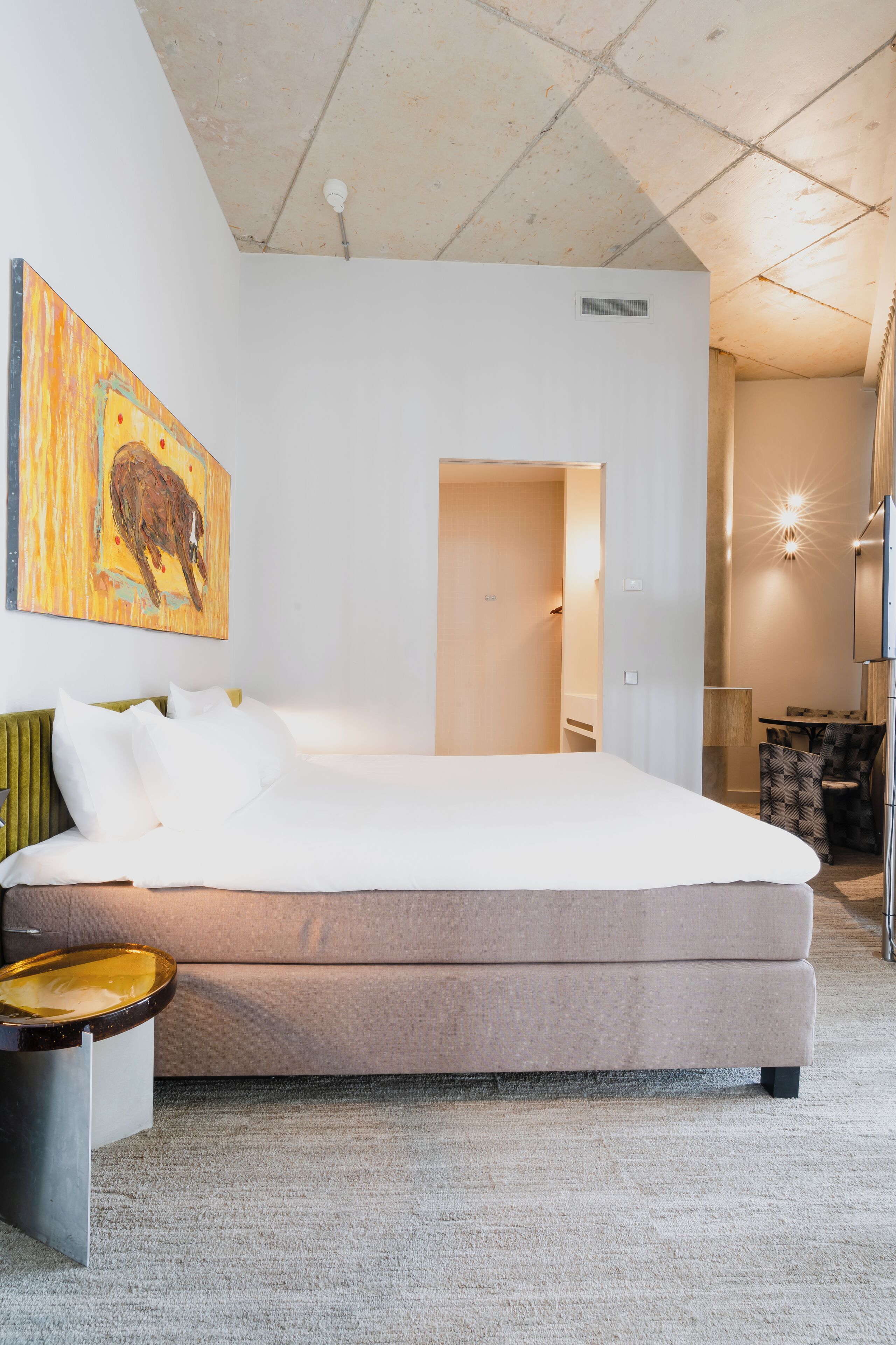 A modern hotel room with a large bed, a painting on the wall, and a small seating area.