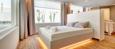Une chambre moderne avec un grand lit, une fenêtre avec des rideaux, un plancher en bois et une petite table avec des serviettes.