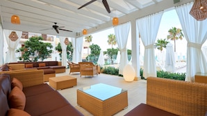 A spacious outdoor lounge area with wicker furniture, ceiling fans, and large windows offering views of palm trees and a pool. - Protur Bonamar Hotel - Adults Only (Son Servera)