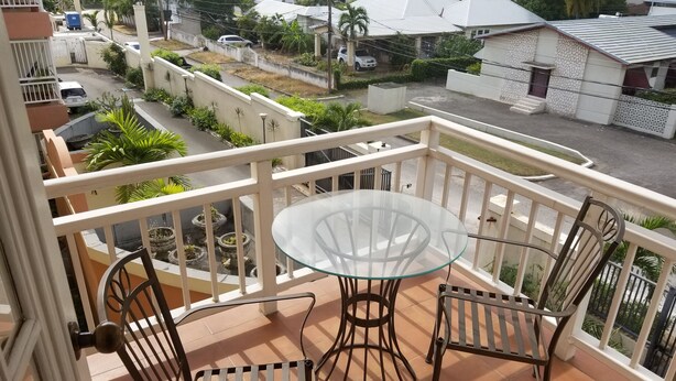En balkon med et glasbord og metalstole, med udsigt over et boligområde med huse og en vej.