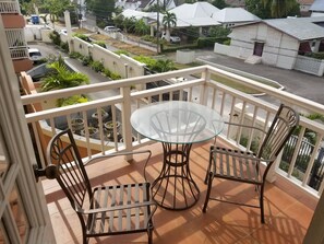 Ein Balkon mit Glastisch und Metallstühlen, der einen Blick auf ein Wohngebiet mit Häusern und einer Straße bietet.
