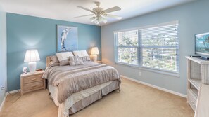 Une chambre à coucher comprenant un lit, des lampes de chevet, un ventilateur de plafond, une fenêtre avec des stores et un téléviseur posé sur un meuble.