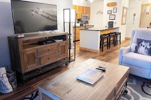 A living room with a TV, wooden cabinet, and a coffee table. There's a kitchen area with wooden cabinets and a microwave. - Great Value Close to Resort w/Heated Pool-Hot Tubs-Grills-Looping Ski Shuttle-Gas Fireplace-Updated (Steamboat Springs)