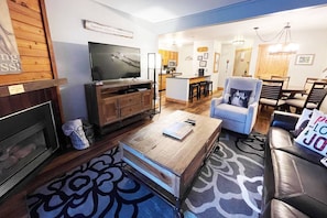 A living room with a TV, wooden entertainment unit, and a fireplace. There is a dining area with a kitchen in the background. - Great Value Close to Resort w/Heated Pool-Hot Tubs-Grills-Looping Ski Shuttle-Gas Fireplace-Updated (Steamboat Springs)