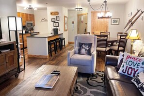 A modern living room with a sofa, armchair, and coffee table. There is a kitchen area with a microwave and bar stools. A dining table with chairs is visible in the background.