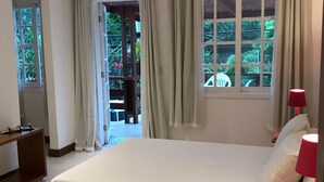 Une chambre avec un grand lit, une table de chevet en bois, un bureau et une chaise. Il y a des fenêtres avec des rideaux, une vue sur la verdure à l’extérieur et une porte qui donne sur un espace extérieur.