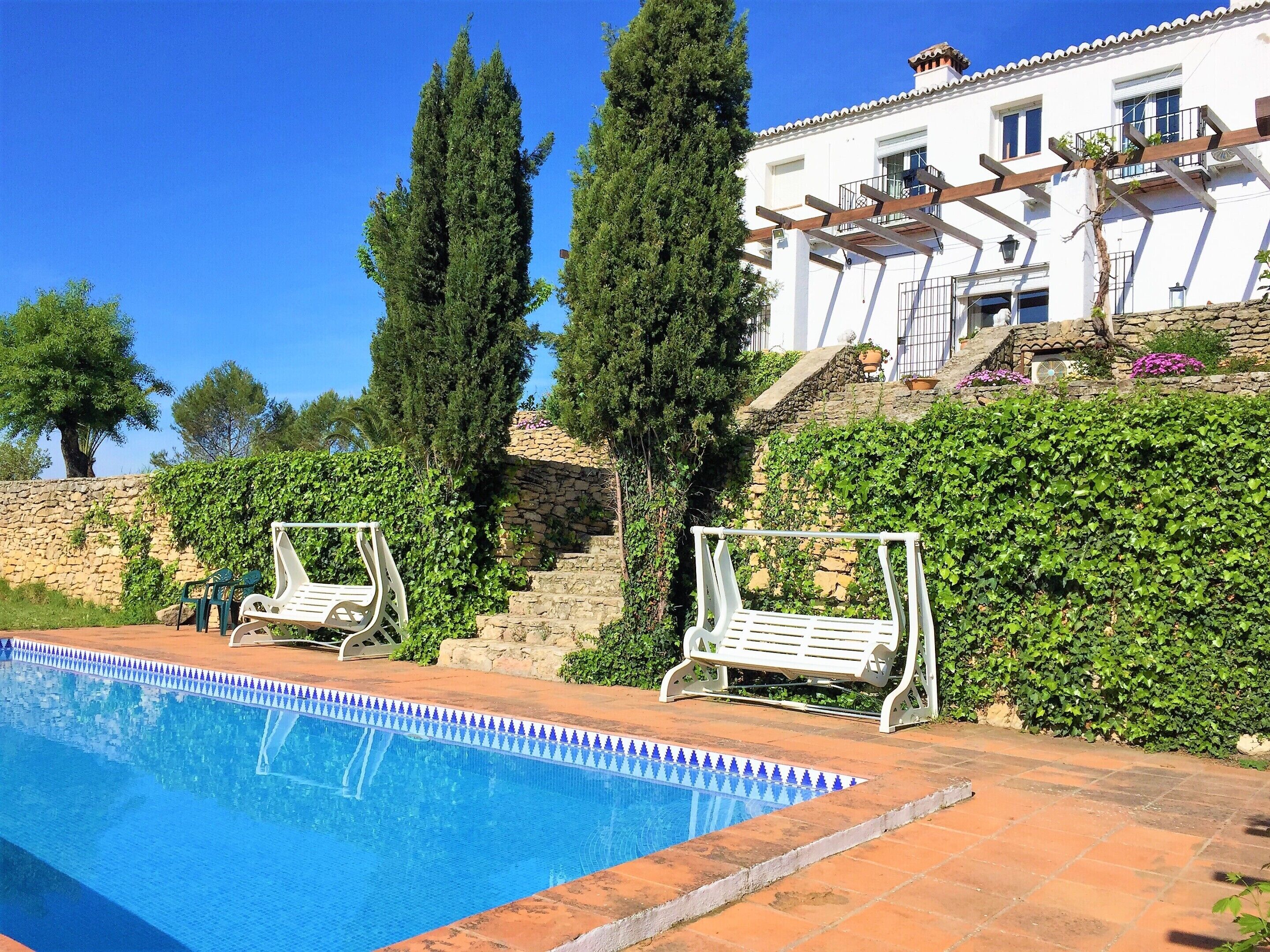 A swimming pool with two white lounge chairs, a stone wall, and a white building with a balcony.