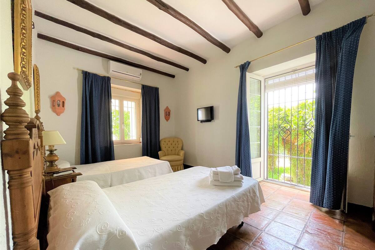 Une chambre avec deux lits, une chaise, un téléviseur et un balcon avec des rideaux.