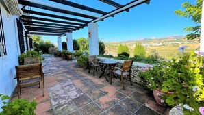 Un patio cubierto con una mesa y sillas, plantas en macetas y vistas al campo.