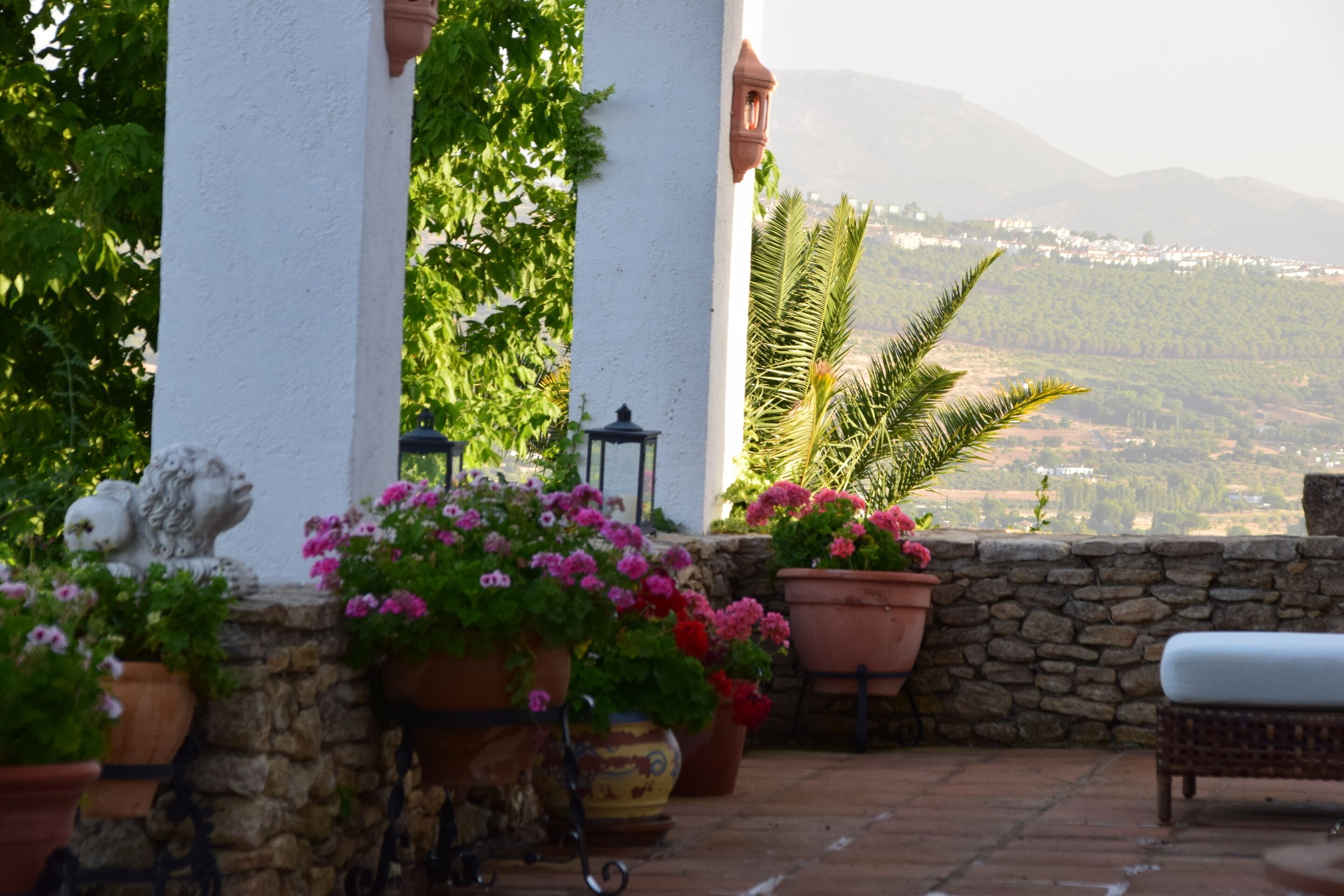 A terrace with potted plants, a stone wall, and a view of a distant town and mountains.