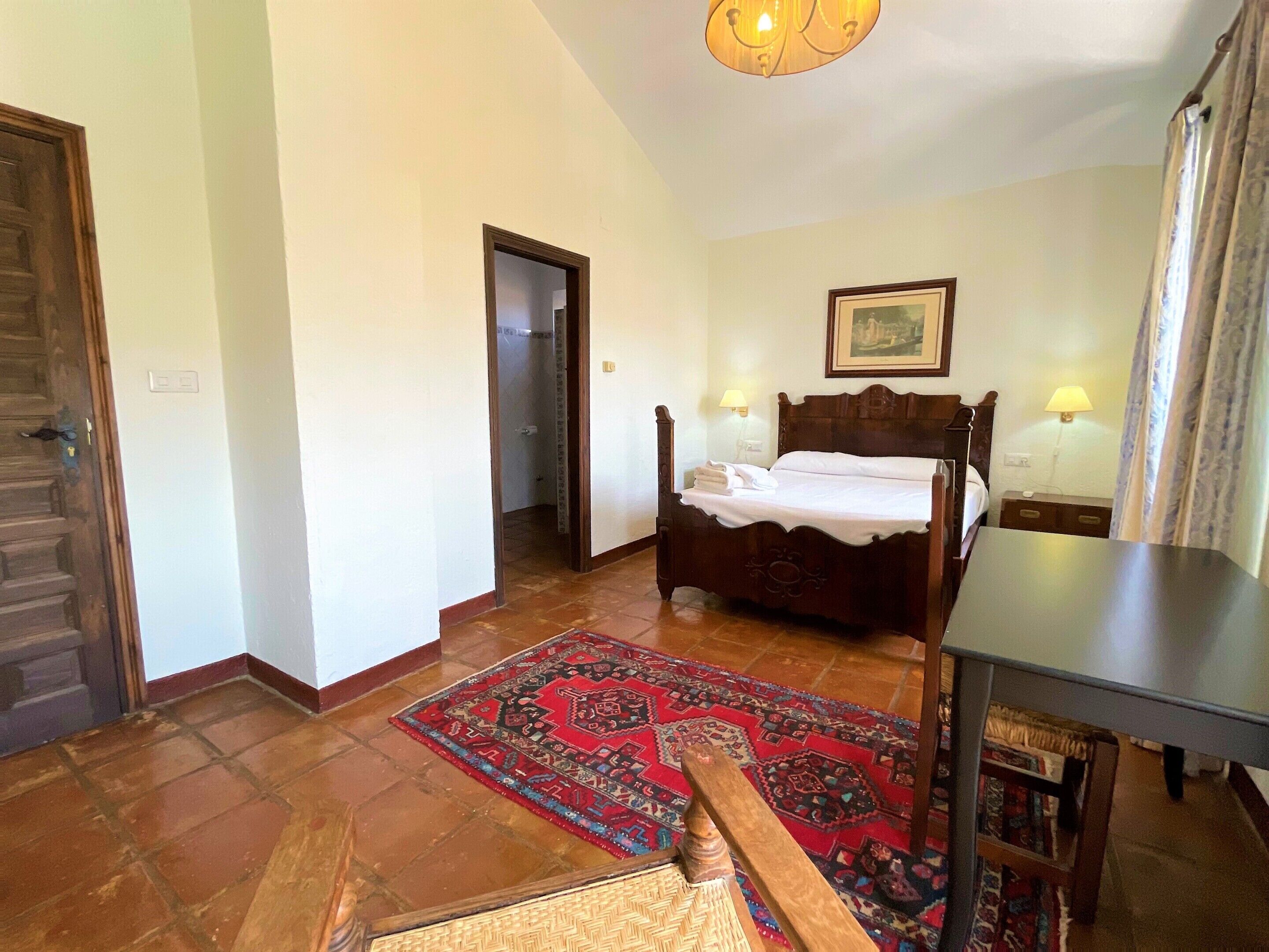 Un dormitorio con cama de madera, dos lámparas de noche, un tapete rojo estampado y un banco de madera.