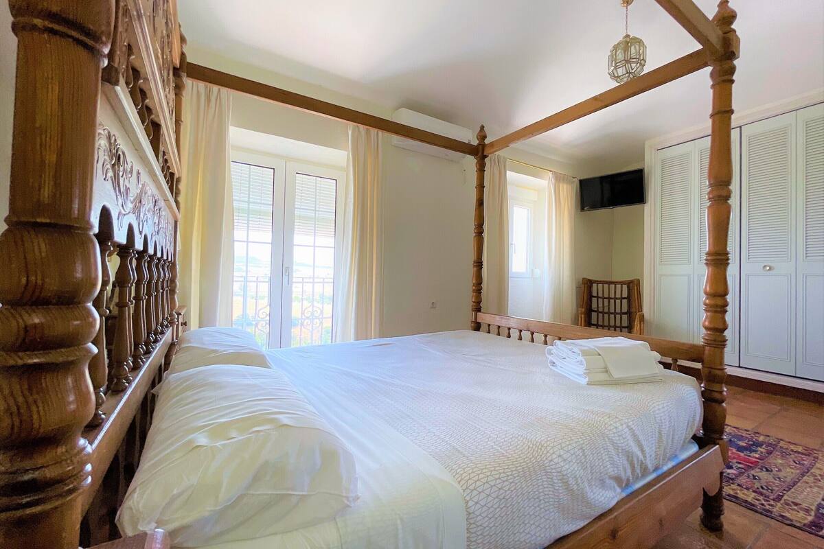 Un lit à baldaquin avec une tête de lit en bois, un téléviseur à écran plat fixé au mur et une grande fenêtre avec des rideaux.