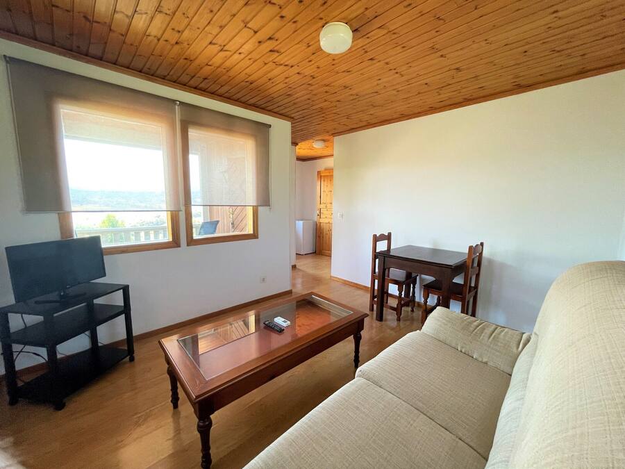 Una sala de estar con sofá, una mesa de centro, televisor y una mesa de comedor con sillas. Hay ventanas con persianas, techo de madera y vistas a un balcón.