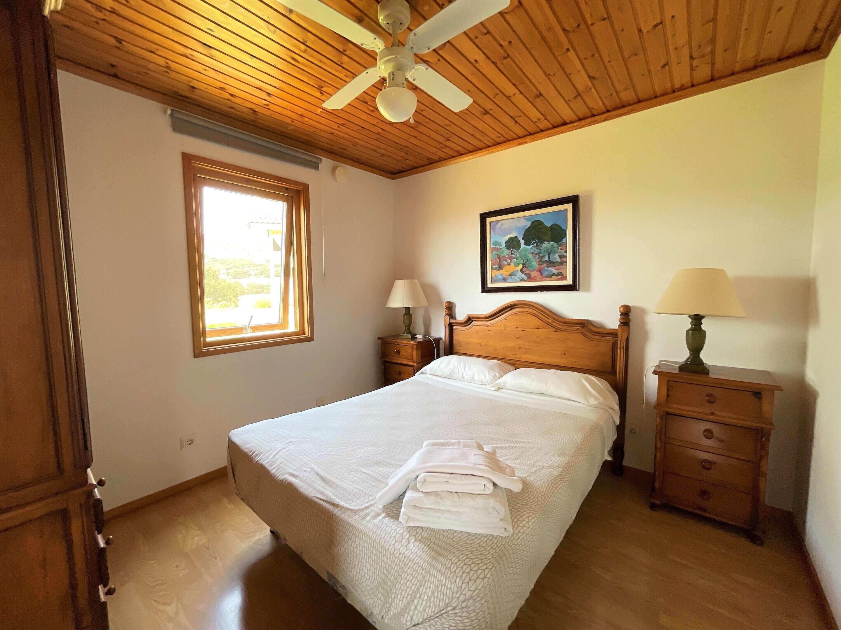 Quarto com teto de madeira, cama com roupa de cama branca, duas mesas de cabeceira com luminárias, uma janela e um quadro na parede.