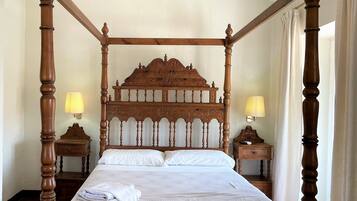 Una cama con dosel, cabecera y piecera de madera, dos mesitas de noche con lámparas, y una cama bien tendida con ropa blanca.