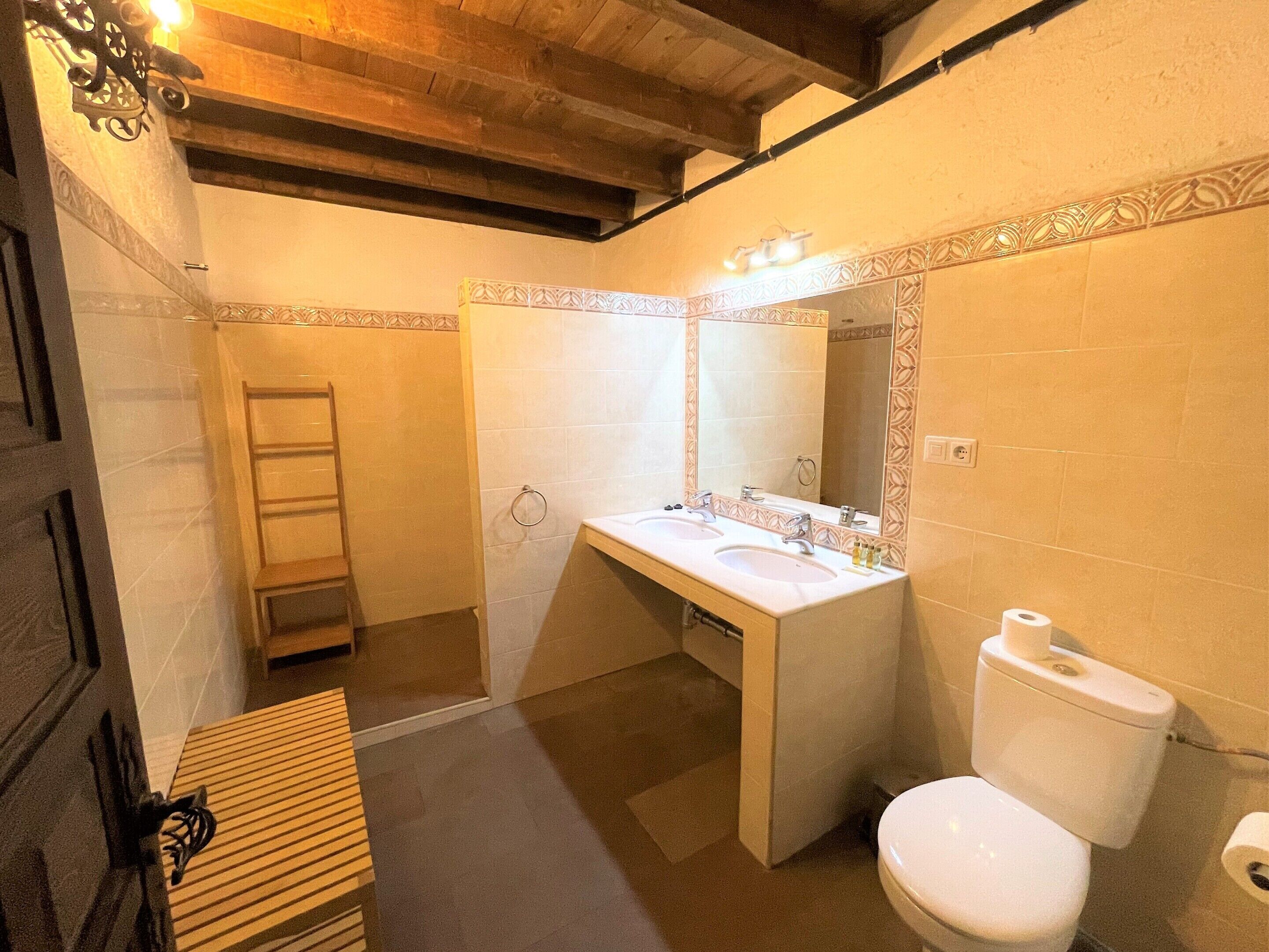 A bathroom with a wooden bench, a double sink vanity, a toilet, and a mirror with lights.