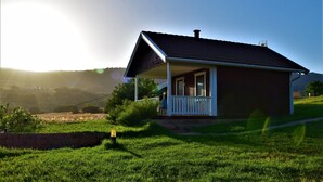 Una casita con un porche, rodeada de un campo de césped y colinas al fondo.