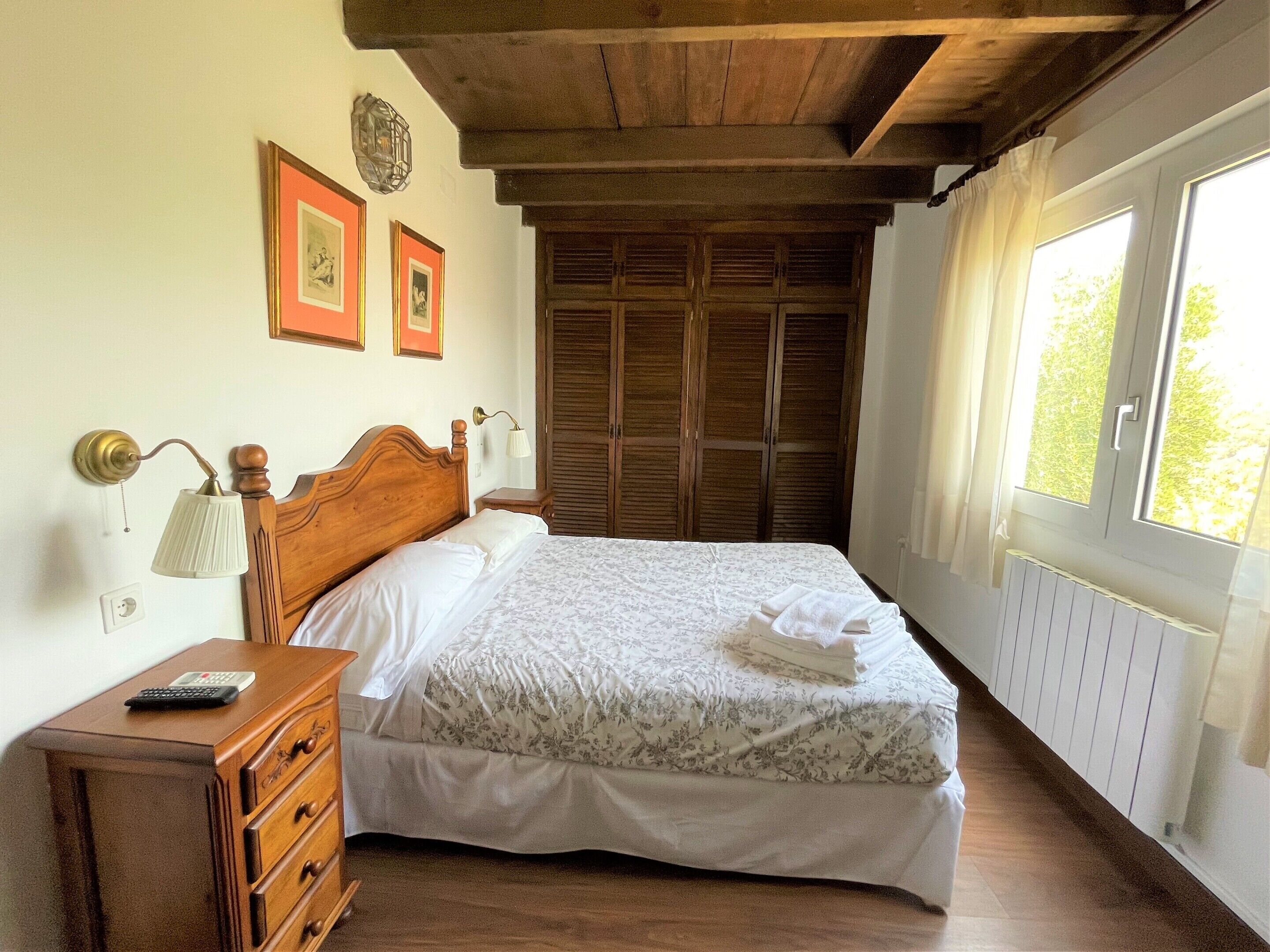 Un dormitorio con una cama de madera, una mesita de noche con lámpara, un aparador y una ventana con cortinas.