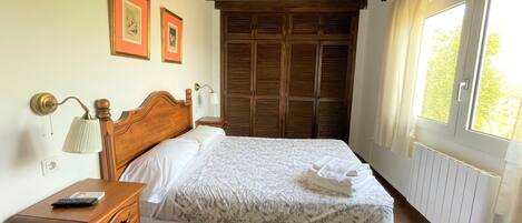 Un dormitorio con una cama de madera, una mesita de noche con lámpara, un aparador y una ventana con cortinas.