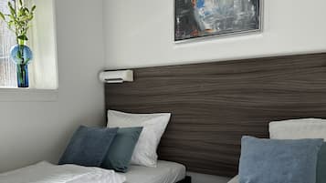 A hotel room with two beds, a window with a plant, a painting on the wall, and a wall-mounted air conditioner.