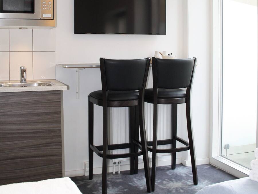 A modern hotel room with a kitchenette, two black bar stools, a flat-screen TV, a bed, and a large window.