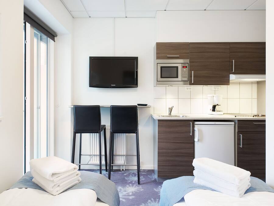 A modern hotel room with a kitchenette, a flat-screen TV, two bar stools, and a bed with towels.