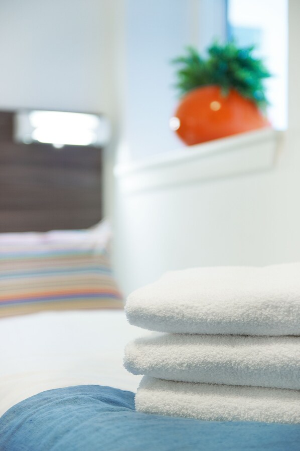 A neatly made bed with a striped rug, a white wall, and a potted plant in the background.