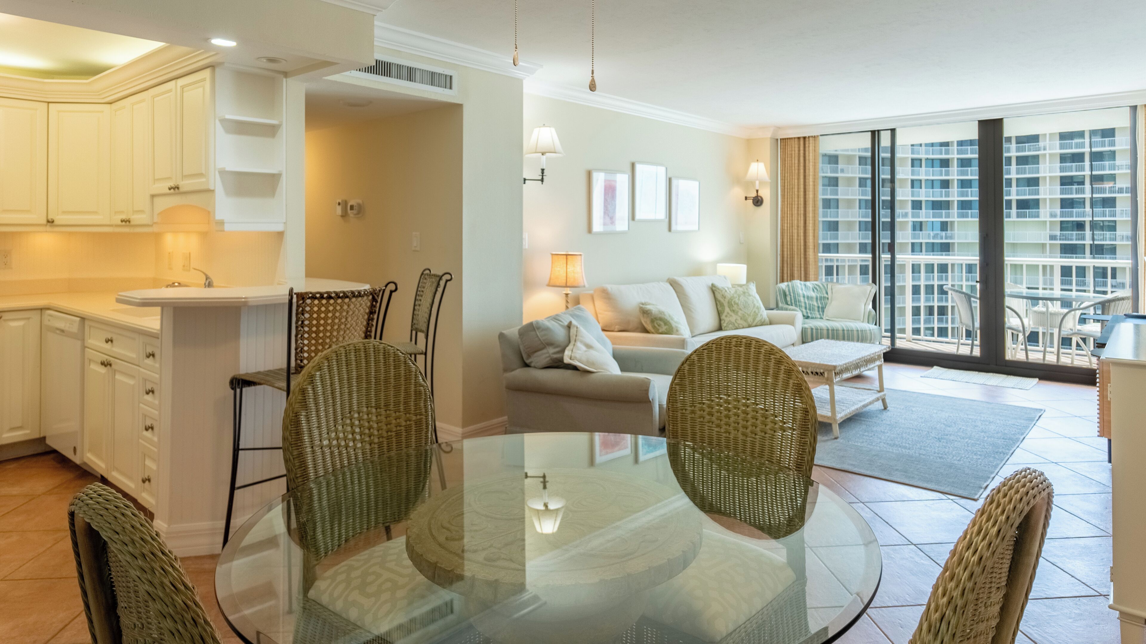 A spacious living area with a glass dining table, wicker chairs, a sofa set, and a view of a high-rise building through large windows.