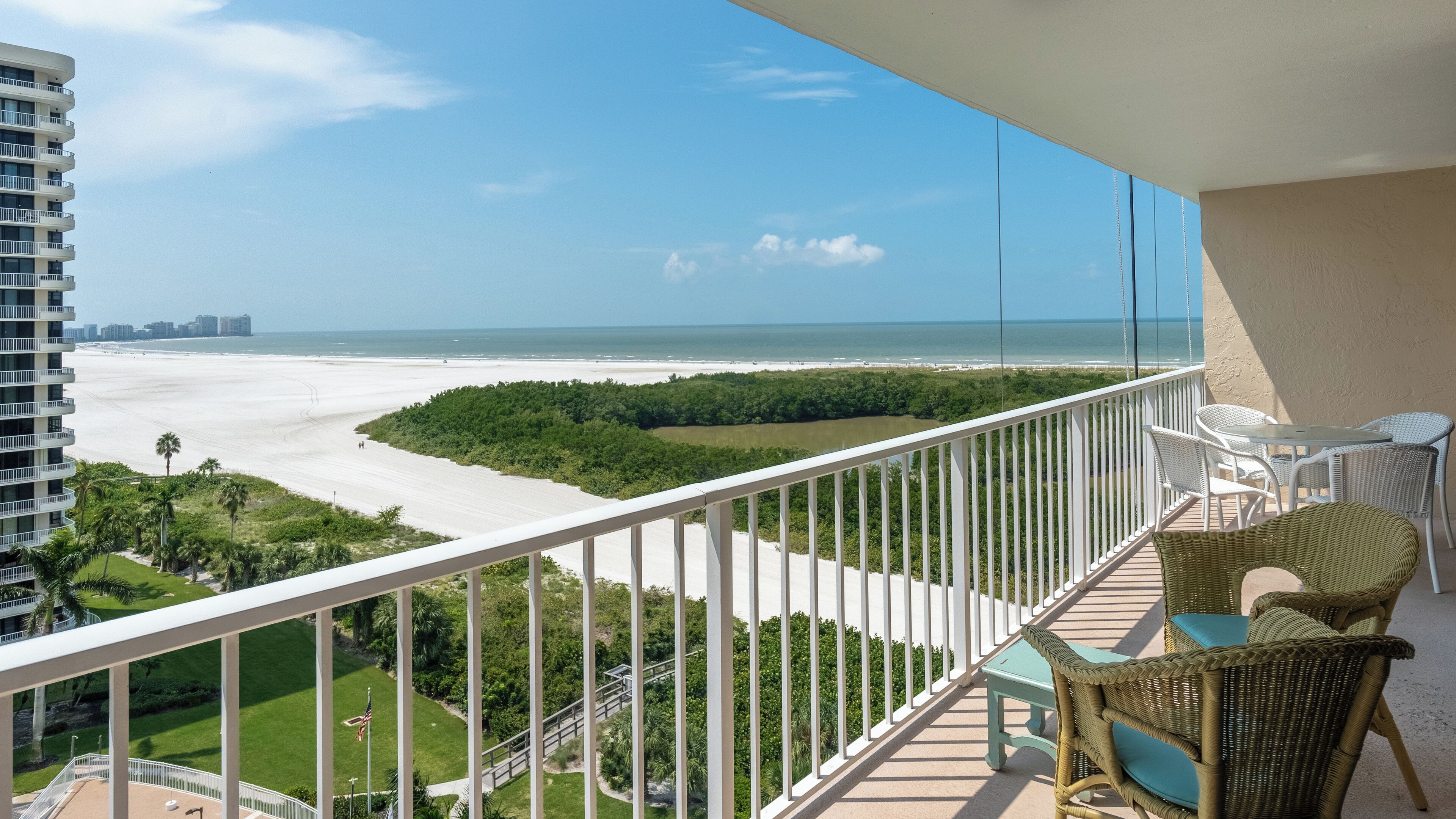 A balcony with a view of a beach, white wicker furniture, and a clear sky.