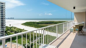 Un balcón con vistas a una playa y al océano, muebles de exterior blancos y una bandera.