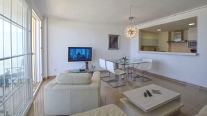 A modern living room with a TV, sofa, dining table, and kitchen area visible through a glass wall. - Harborfront Escape in Mallorca (Ca'n Picafort)
