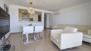 A modern living room with a white sofa, a dining area with a white table and chairs, and a large mirror above the counter. - Harborfront Escape in Mallorca (Ca'n Picafort)