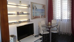 Un salon moderne avec un téléviseur à écran plat, une table à manger en verre, des chaises blanches, un tableau représentant un paysage côtier et un rideau rouge.