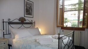 Un lit bien fait, avec du linge de lit blanc, une table de chevet avec une lampe et une fenêtre donnant sur des bâtiments et des arbres.