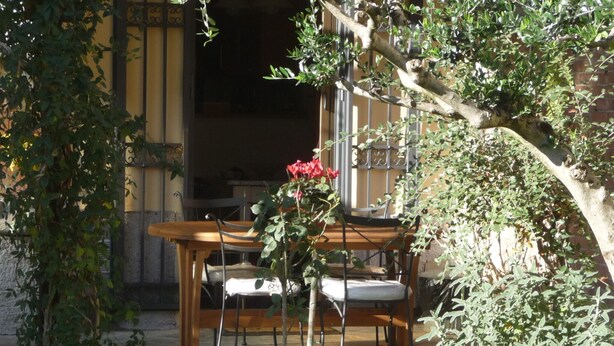 Une table et des chaises en bois sont disposées à l’extérieur, entourées de verdure et d’un bâtiment doté d’un portail en métal.
