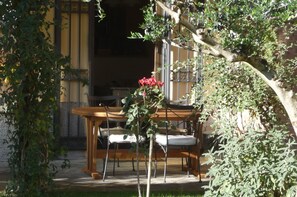 Conjunto de mesa e cadeiras de madeira ao ar livre, rodeado por vegetação e um edifício com portão de metal.