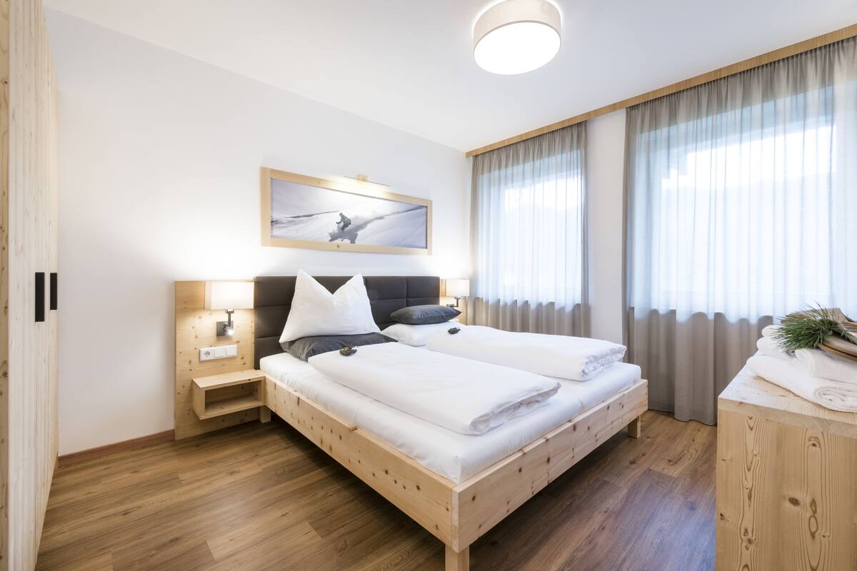 Ein modernes Hotelzimmer mit einem Holzbett, einem Nachttisch, einem Schreibtisch und einem großen Fenster mit Vorhängen.