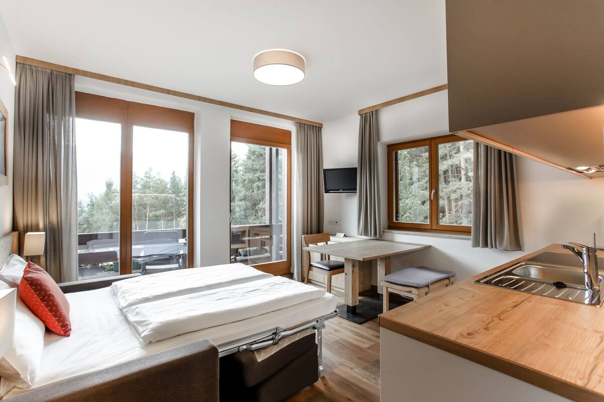 Ein modernes Hotelzimmer mit Bett, Schreibtisch, Sessel, Fernseher und einer Küchenzeile mit Spüle und Arbeitsfläche.