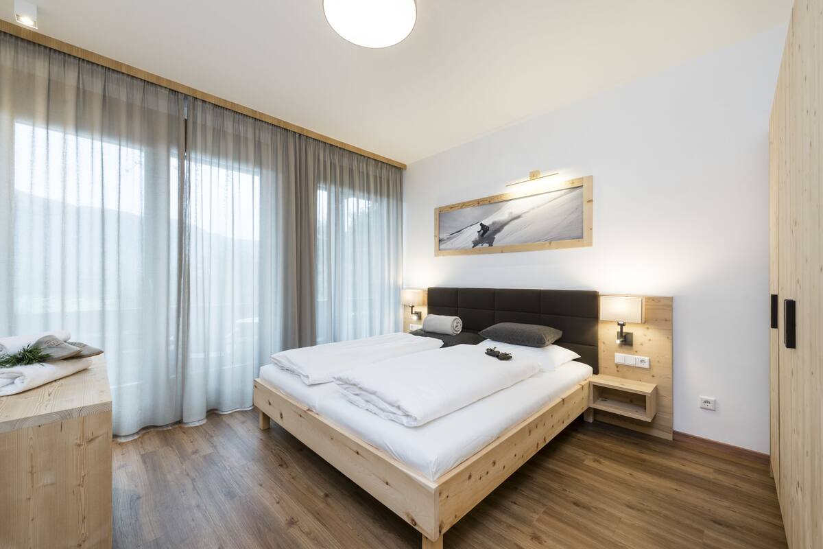 Ein modernes Schlafzimmer mit einem hölzernen Bett, einem großen Fenster mit durchscheinenden Vorhängen, einem aufgehängten Gemälde und einem Nachttisch mit einer Lampe.