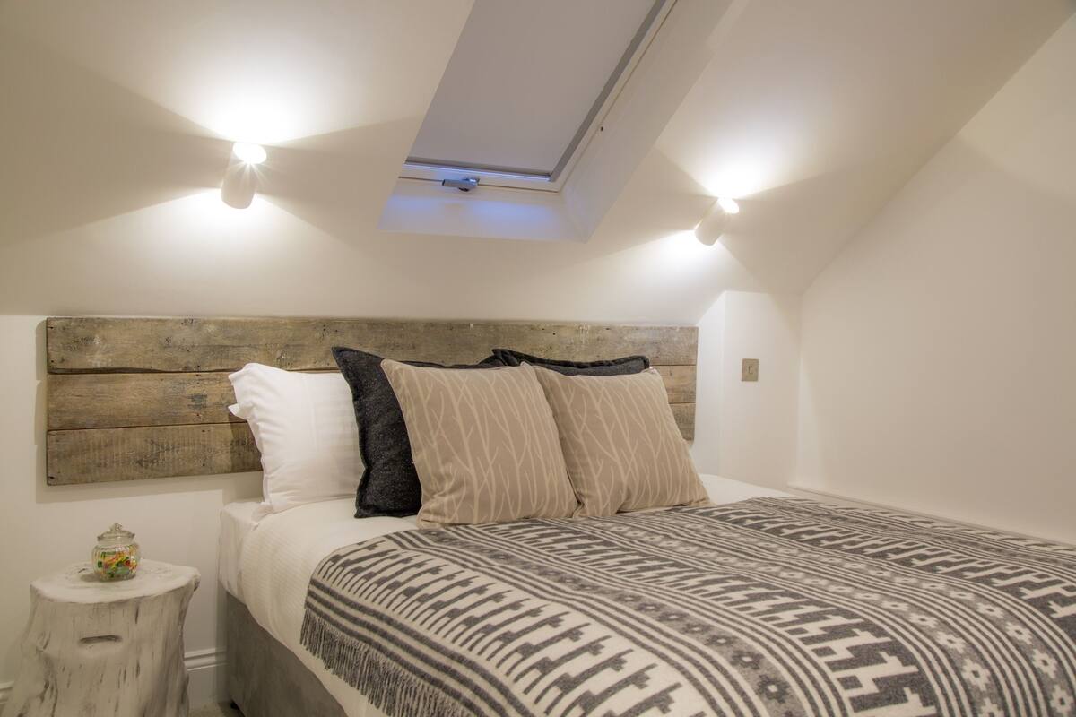 A bedroom with a sloped ceiling, a wooden headboard, a patterned bedspread, and two wall-mounted lights.