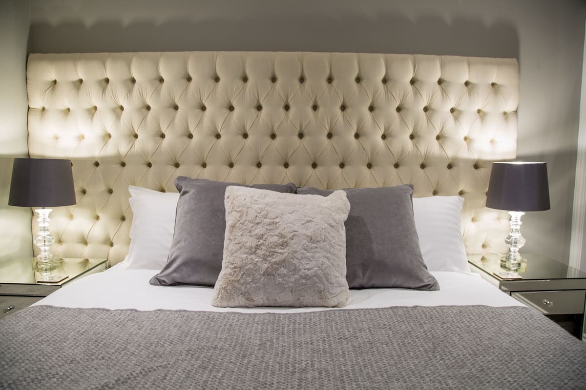 A bedroom with a tufted headboard, a bed with white and gray bedding, two bedside lamps, and a glass side table.
