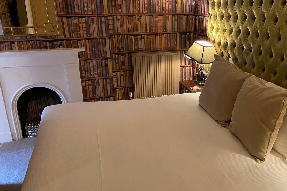 A large bed with a tufted headboard, a fireplace, and a bookshelf in the background.