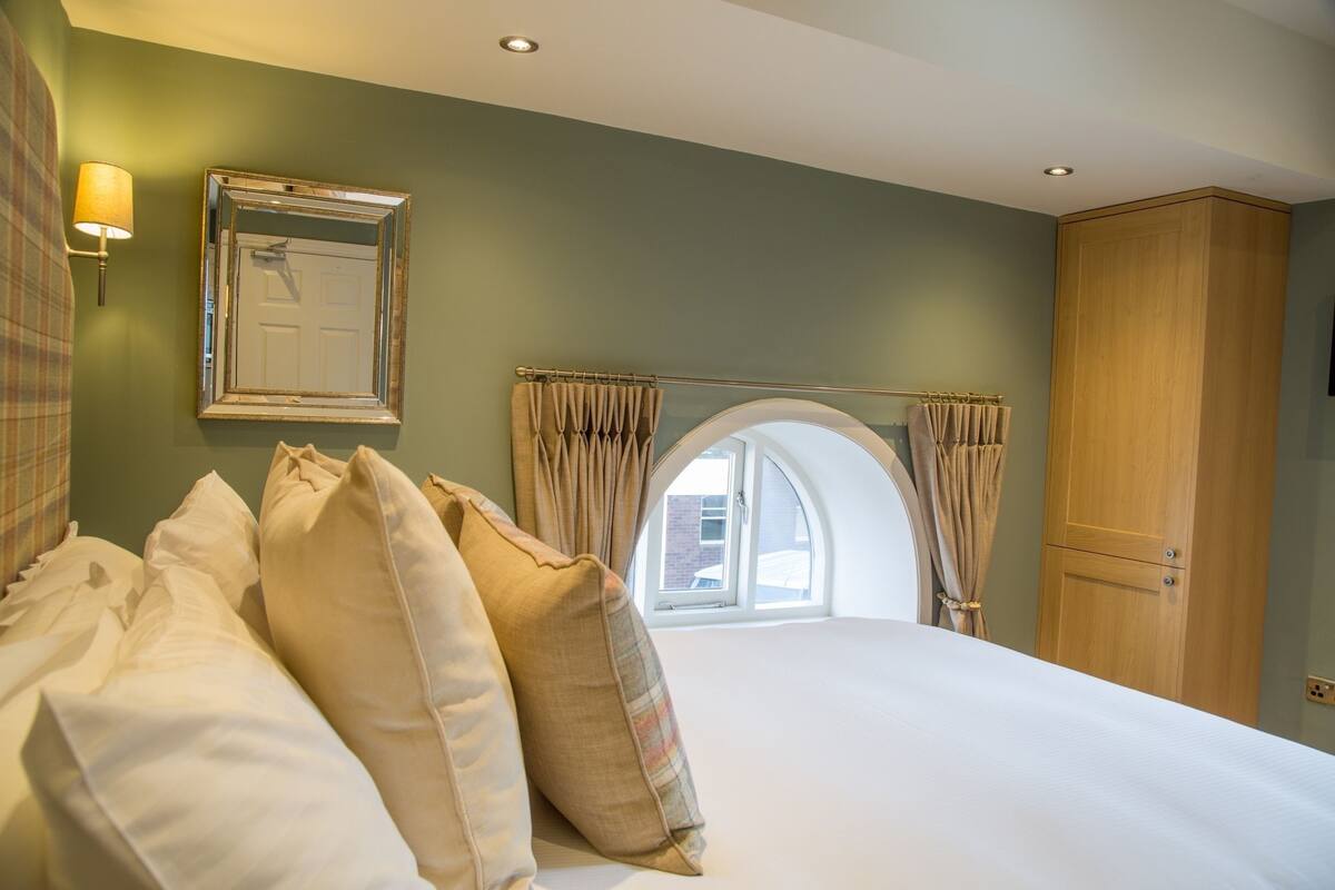 A neatly made bed with pillows, a mirror, and a wardrobe in a room with green walls and a window.