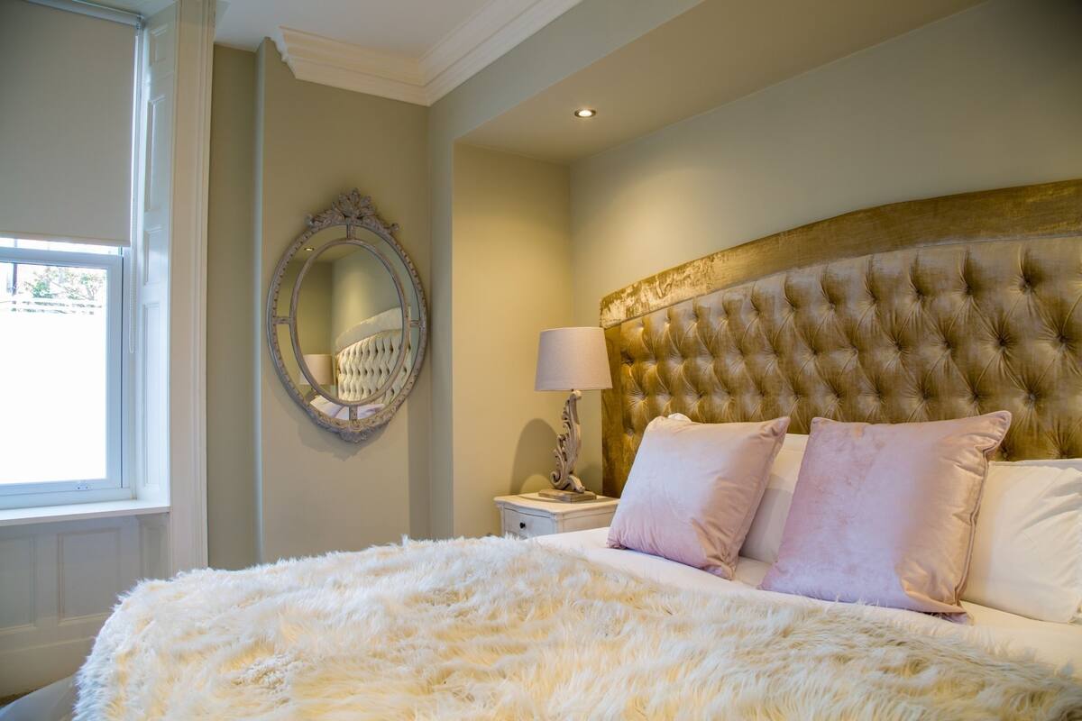 A bedroom with a tufted headboard, a fluffy white bedspread, and a round mirror on the wall.