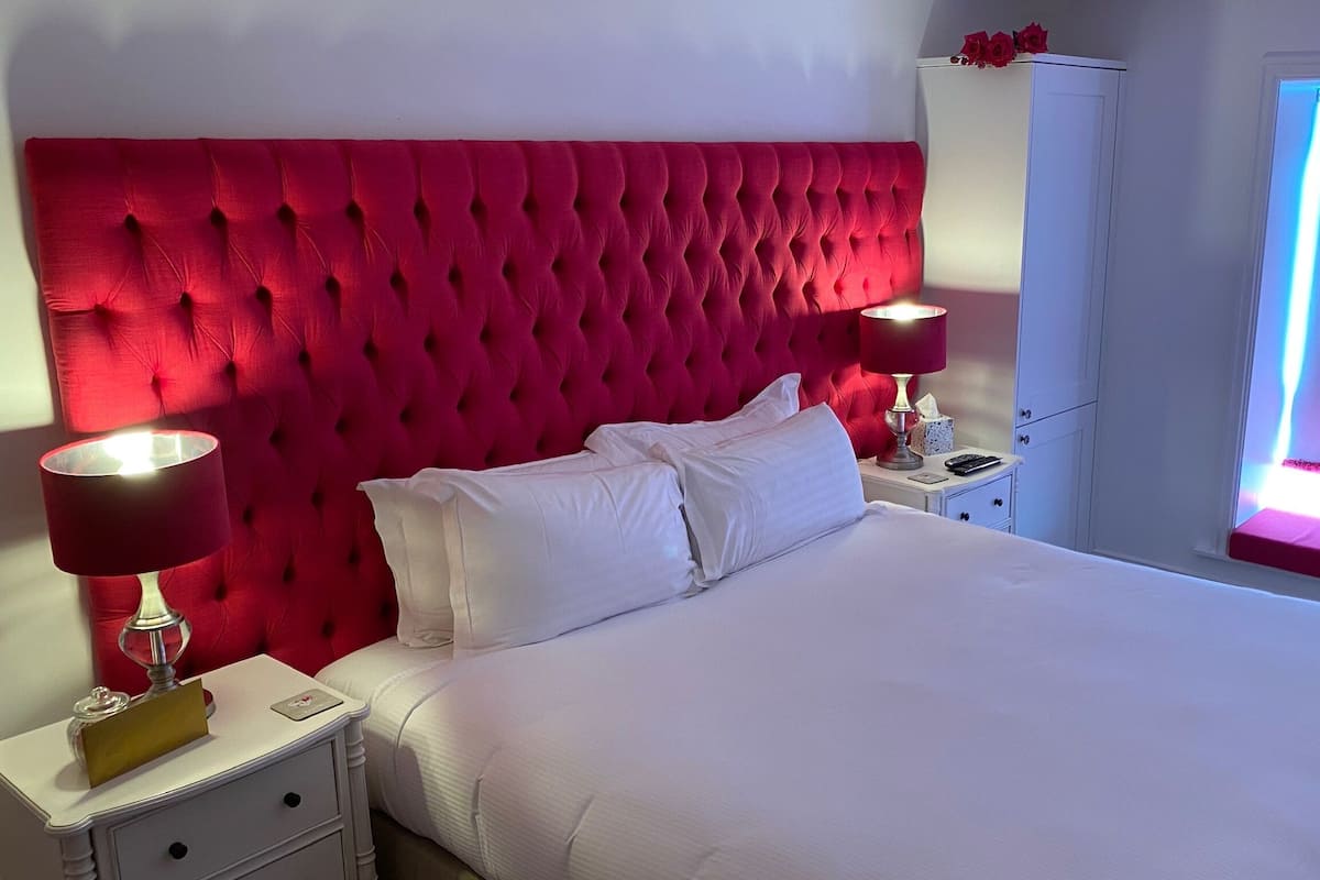A bedroom with a tufted red headboard, white bedding, a bedside table with a lamp, and a window with a pink light.