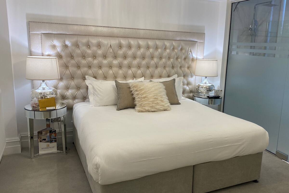 A bedroom with a tufted headboard, a bed with white bedding, two bedside tables with lamps, and a glass-enclosed wardrobe.