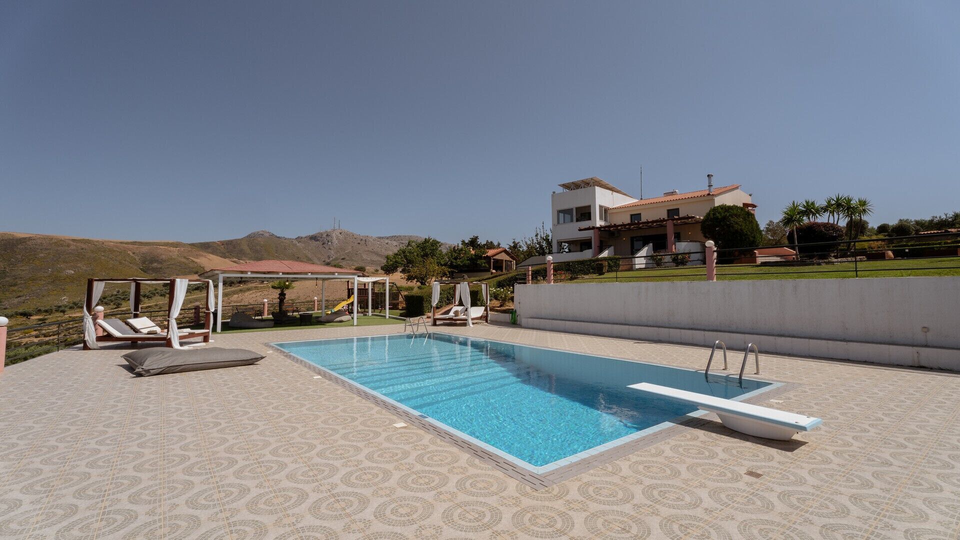 Ein Swimmingpool mit Liegestühlen und Sonnensegel, umgeben von einem gemusterten Boden, mit einem Gebäude und Hügeln im Hintergrund.