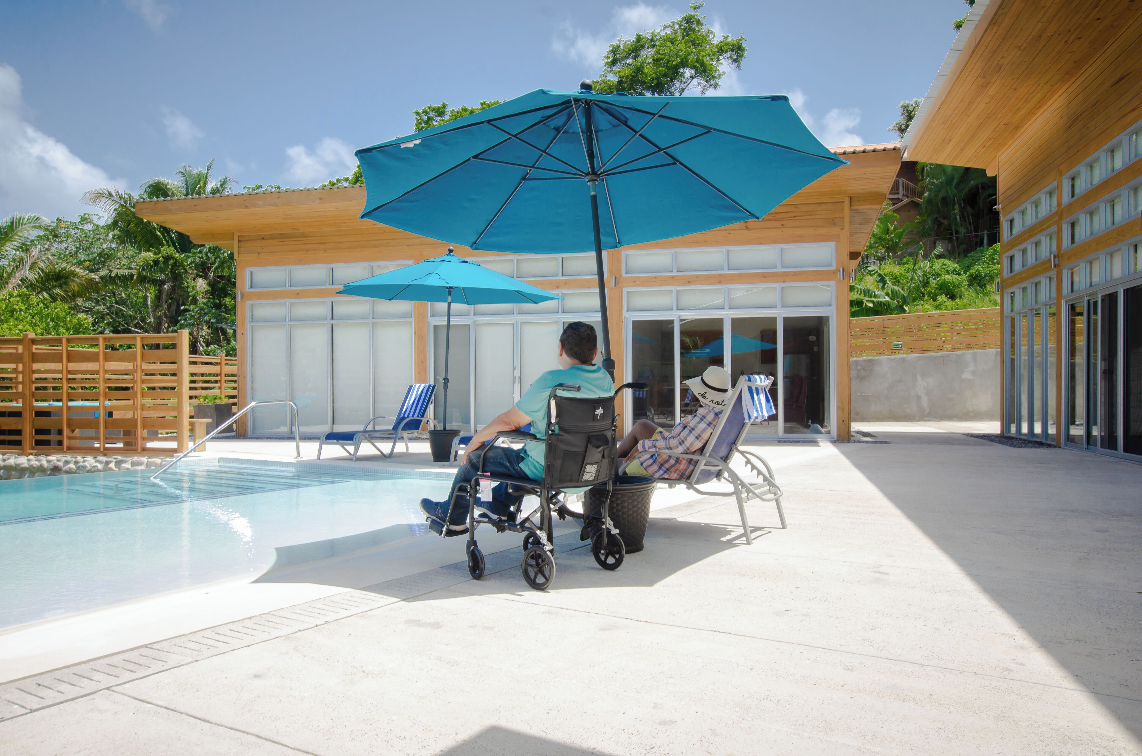 Zona de piscina con una silla de ruedas, una persona en silla de ruedas y otra persona sentada en una tumbona.