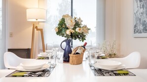 Una mesa de comedor con platos, tazones y cubiertos blancos, un florero con flores, una botella de vino y una lámpara.